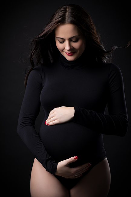 babybauch Fotografie in Düsseldorf. Schwangere in schwarz gekleidet. Blick nach unten auf ihren Bauch gerichtet, den sie mit ihren Händen hält.