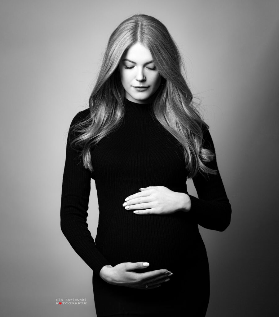Babybauchshooting Düsseldorf. Schwangere Schönheit in schwarzem Kleid hält zärtlich ihren Schwangerschaftsbauch und senkt ihren Blick nach unten.  